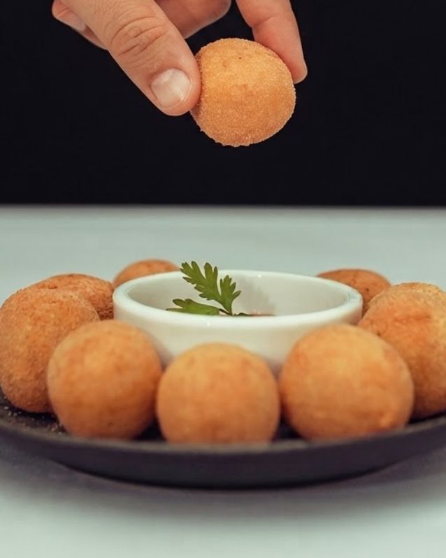 Mão pegando um bolinho de charque com banana de um prato com outros bolinhos e molho