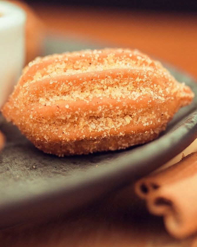 Churros com açúcar num prato escuro, com pote de doce de leite e canela ao lado